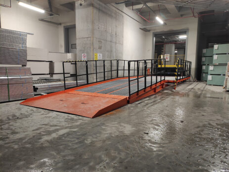 Industrijska notranja uporaba, kjer so nameščene oranžne nakladalne rampe z zaščitnimi ograjami, namenjene učinkovitemu pretovarjanju tovora v skladišču.