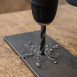 Akumulatorski vrtalnik s svedrom, ki vrta skozi kovinsko ploščo in ustvarja spiralne kovinske ostružke – prikaz delovanja, kot ga omogočajo najboljši svedri za kovino.